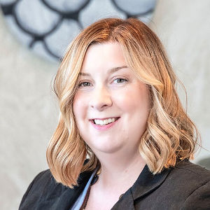 A woman with shoulder-length brown hair and a fuchsia pink shirt smiles at the camera. She has light skin and hazel eyes.
